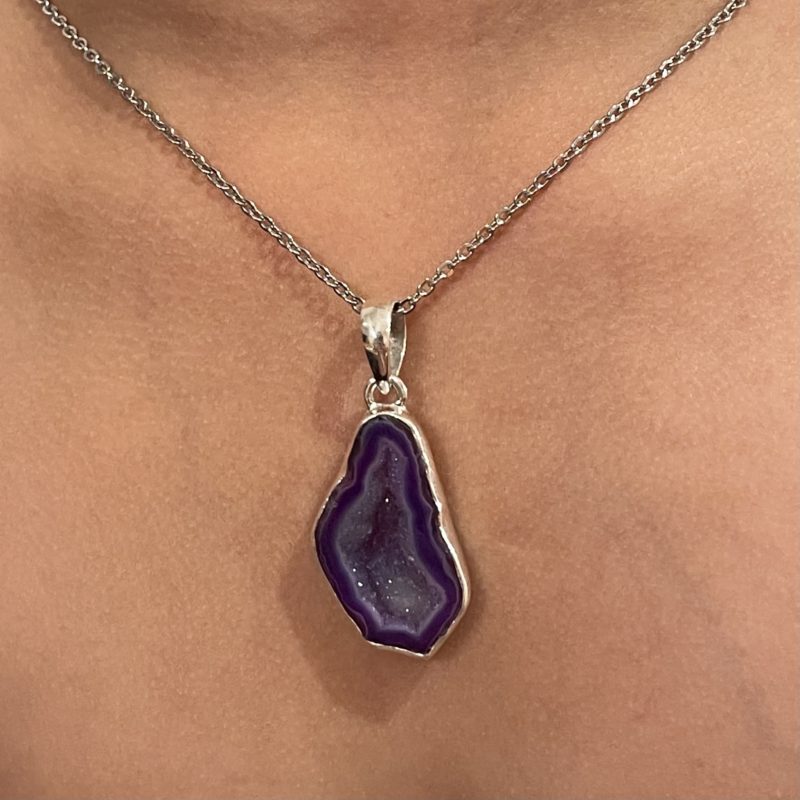 An Agate Necklace adorned with a purple druzy pendant gracefully rests on a woman's neck.