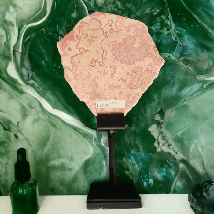 A Rhodochrosite Slice On Stand 300mm high next to a green marble wall.